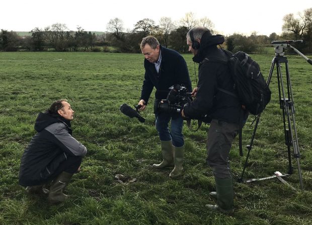 A farmer kneeling in a field while being interviewed by a TV crew