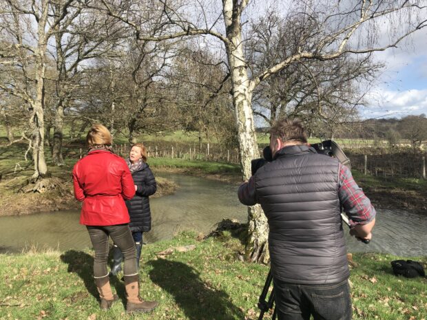 A TV cameraman with his back to the camera in front of an interview with a Natural England by a pond