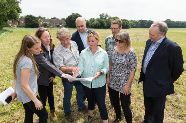 An image of several people standing in a green field looking at some paper plans.
