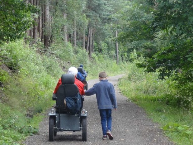 A young children and elderly man in a wheelchair walking down a country lane 