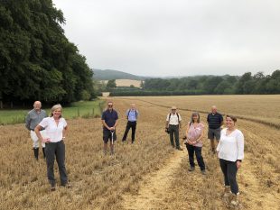 A group of people are standing socially distanced in a wheat field. There are trees in the background and rolling hills. 