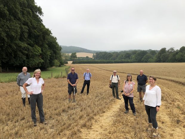 A group of people are standing socially distanced in a wheat field. There are trees in the background and rolling hills.