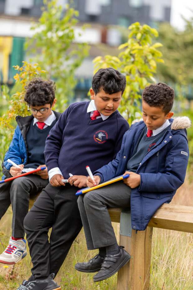 three boys sit on a bench in nature, with their clip boards writing 