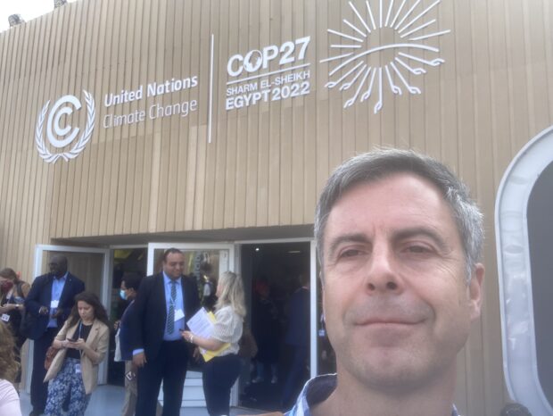 A selfie picture of Mike, stood outside the COP27 centre. There are many people in the background and a large COP27 sign behind him.