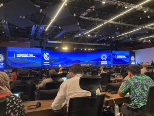 Mike is sat in a very busy, dark room. There are many people sat at desks listening to discussions from speakers. There is a large blue panel light above at the front of the room.