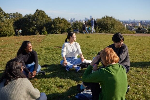 A group of young people of all backgrounds, sit together in a circle, laughing and talking. They are within an urban park, surrounded by a green field. There are trees in the distance, and a cityscape on the horizon 