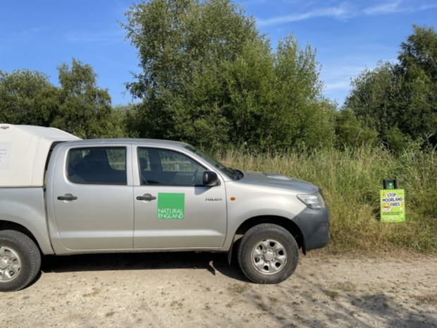 A silver pick up truck is parked at the nature reserve. The truck has a green Natural England logo on the side. 