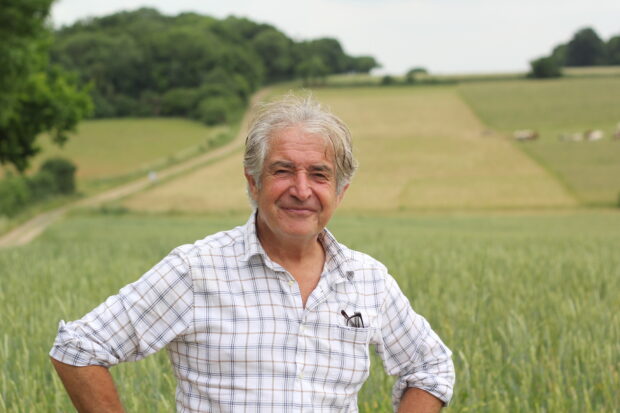 Photo shows Tony Juniper, Chair of Natural England, stood within a green open field. He wears a white checked shirt and stands with his hands on hips. There are trees in the distance.