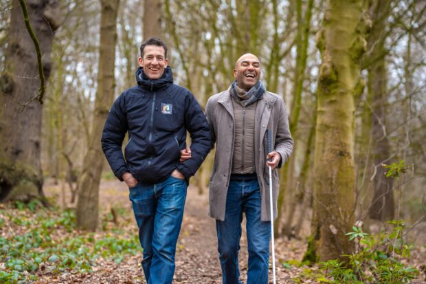 Amar Latif, President of the Ramblers and blind adventurer is walking in the woods with a colleague. Amar is wearing a brown overcoat and is carrying a white cane. He is smiling broadly. His colleague is wearing a blue rain jacket and has his hands in his jean pockets. He is also smiling. The trees in the woods don’t have any leaves and it appears to be a chilly day.