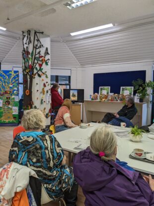 Photo of a group of people at one the pilots in Hampshire. The group are sat within a library room which has lots of nature decorations created by children. They are watching a screen and presentation. 