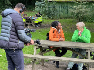 In the image, Shanequa Paris sits with Debbie North on a picnic bench as they discuss the Code. A camera man stands before them filming.