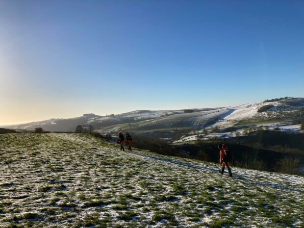 The team climb a tall grassy hill. There is fallen snow or frost coating the ground. A blue sky hangs overhead, and rolling hills in the distance, all covered in white coating.