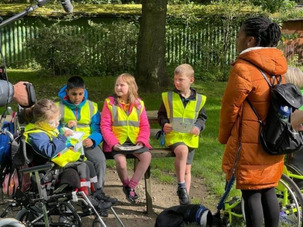 In the image, CBBC Newsround presenter, Shanequa Paris, stands in front of a small group of primary school children, who are sitting on a bench. One child is sat within a wheelchair. The children wear high visibility jackets. Around them, the camera and sound crew hold cameras and boom mics. All are within a small park and discussing the Countryside Code.
