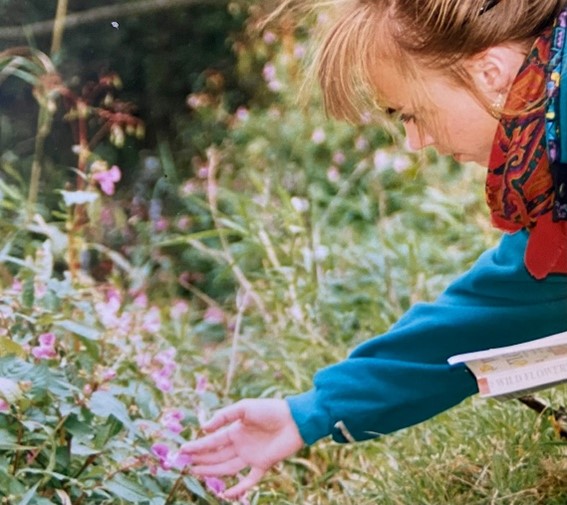 Image shows Clare Warburton when she was younger leaning over to touch some pink flowers