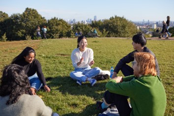 Image shows a group of young people sat in a circle in an urban park. There's a large city in the background