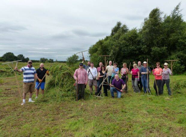 Image shows a group of volunteers standing for a photo op whilst working on a grassy field. There is a bundle of cut vegetation beside them, they carry tools in their hands .