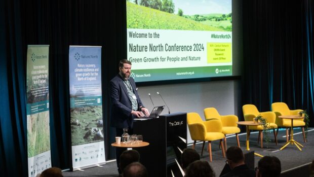 Image shows David Renwick – Regional Director, North for Natural England stood at the front of a conference hall, speaking at a podium. There are empty yellow chairs to his left and a large projector screen displaying a presentation above him.