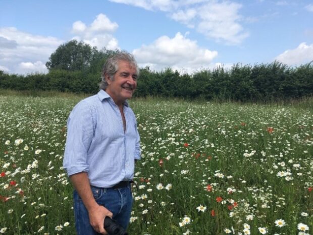 Tony Juniper stands in a grassy field with lots of colourful wildflowers growing. A blue sky with white fluffy clouds hangs overhead.