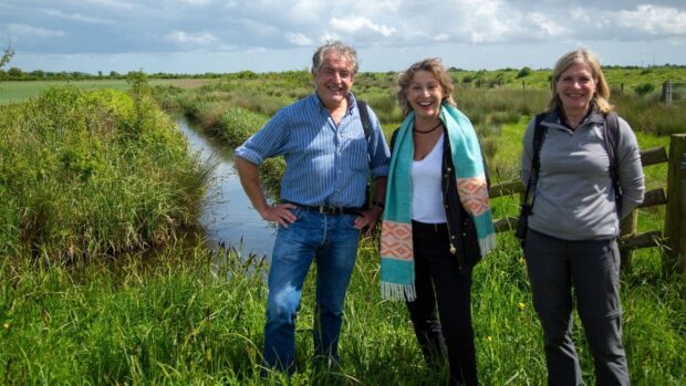 Tony stand with Minister Pow and Marian Spain for a photograph in front of the wetlands. The vegetation is lush and green. A cloudy sky hangs over them. 