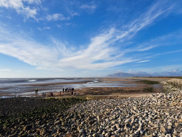 Image shows Earnse Bay, a bright blue sky hangs overhead, with wispy clouds stretching into the distance. A brown sandy shore can be seen for miles. A group of people are stood in the distance, exploring the sand and dunes.