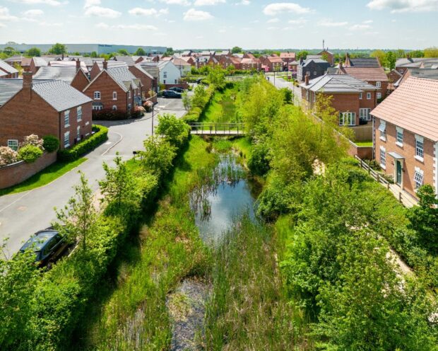 Image shows a lush green canal/waterway running through the centre of a neighbourhood. Red bricked houses stand either side of the new development estate. Lots of trees and bushes grow beside it.