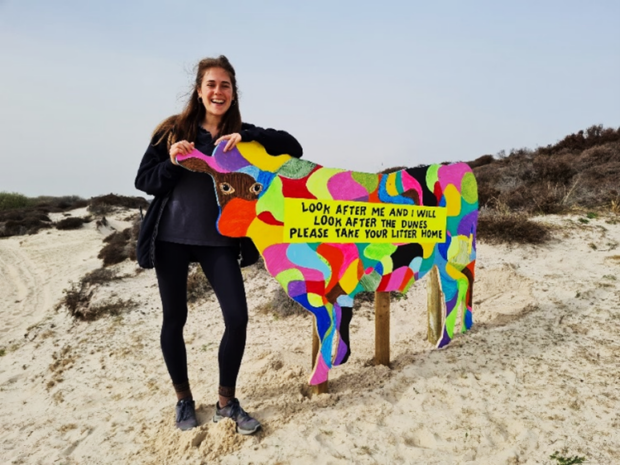 Image shows one of the project leads stood beside an arty cow sign at the dune. The sign is shaped like a cow and is covered in brightly coloured patches of every colour. 