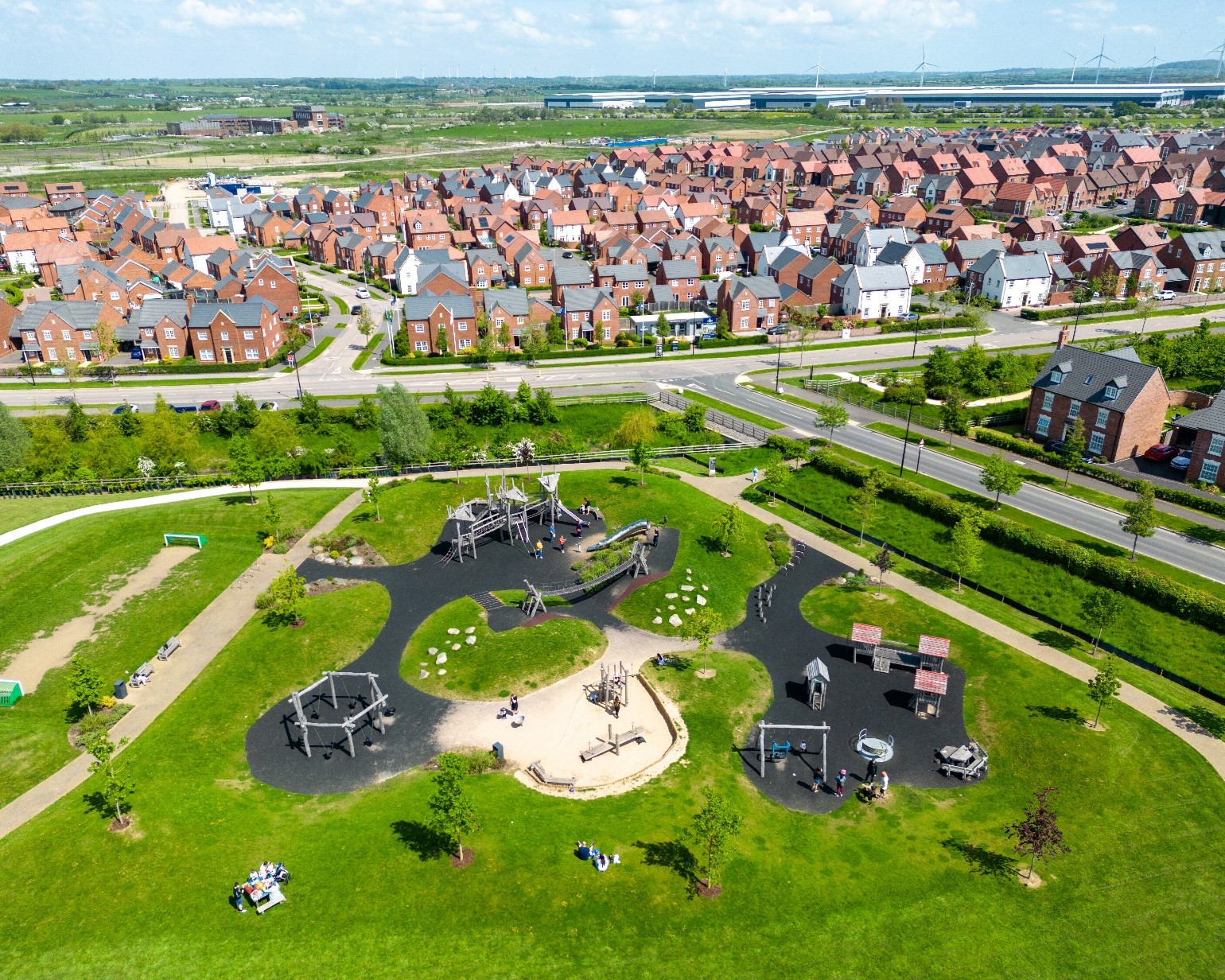 Image shows an aerial view of Houlton housing development, Rugby in the background of the image. In the foreground lies a large green park complete with children's playground. There are trees and bushes planted, dividing the road and park. The area looks lush and green. 