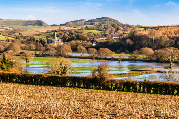 Image shows the village of Otterton, which sits on green hills surrounded by browning trees. A blue sky hangs overhead. Below the village in the foreground, fields are covered in water from the swollen river that is out of shot. The picture shows how the lower ground is easily flooded during heavy periods of rain etc. 