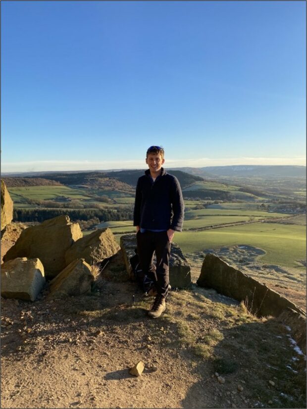 a happy looking geologist under a blue sky, with a green rolling landscape beyond. There are large blocks of sandstone around him.