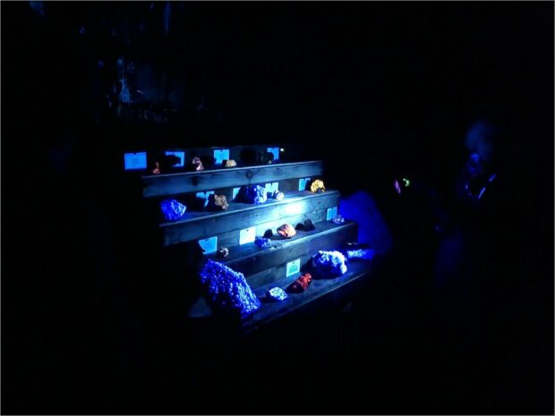 a display shelf is light by a single bluish light, otherwise the picture is in darkness. On the display shelf, minerals glow bright colours, most striking are the yellow and red rocks that are clearly not simply reflecting the blue light