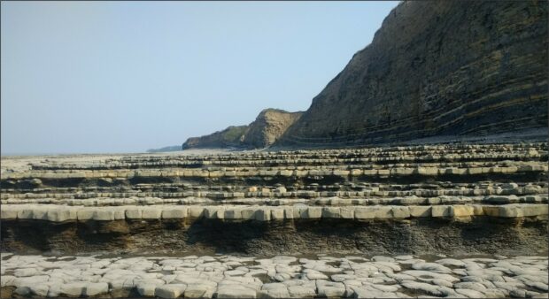 A striking pattern of alternating layers of pale limestone and dark shale. A cliff rises behind the foreshore exposures.