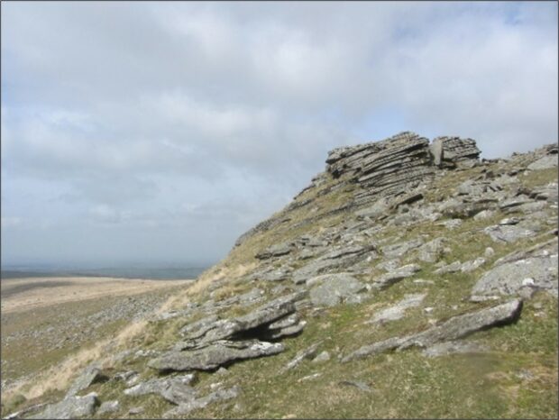 A view across a rocky hillside, with low winter grasses. A fractured granite outcrop (a Tor) dominates the skyline.