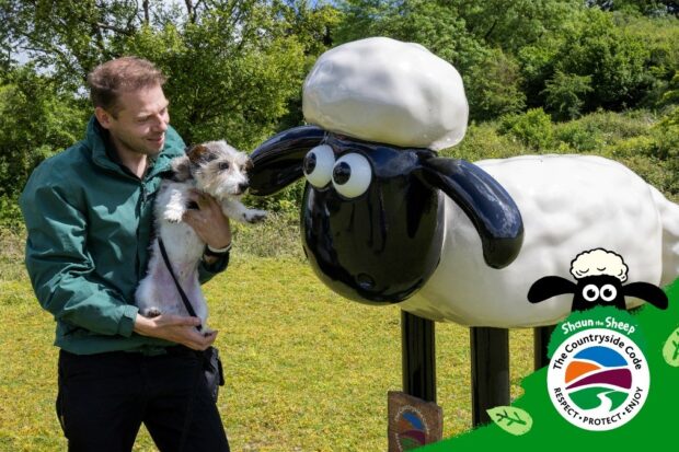 Image shows Tim and Roo from Stokenchurch Dog Rescue with The Countryside Code Champion, Shaun the Sheep statue. Tim hold Roo in his arms whilst they look at Shaun in a grassy field. 