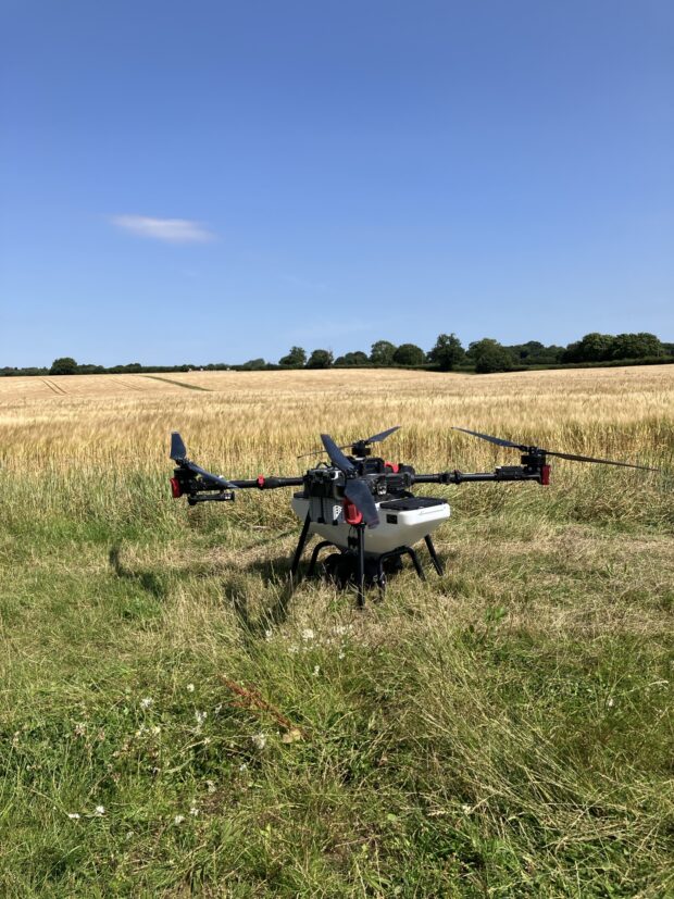 A very large drone, stands ready to fly over the grassy fields to sow seeds. The sky is clear and blue. There are trees in the distance.  Behind the drone is a field of tall golden grain  