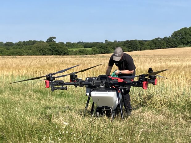 An staff member is preparing a very large drone, ready to fly over the grassy fields to sow seeds. He is stood within a large field, the sky is clear and blue. There are trees in the distance.  