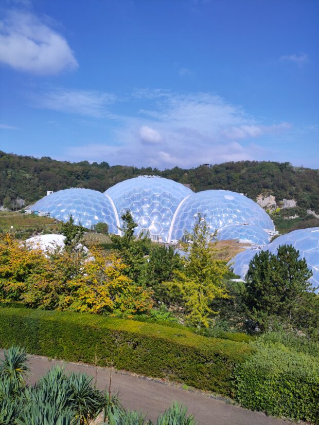 Image shows a picture of  The Eden Centre, a blue sky hangs overhead with fluffy clouds. Lots of green trees are in the foreground, their leaves are just starting to change colour, indicating that it's likely later summer/early autumn time 