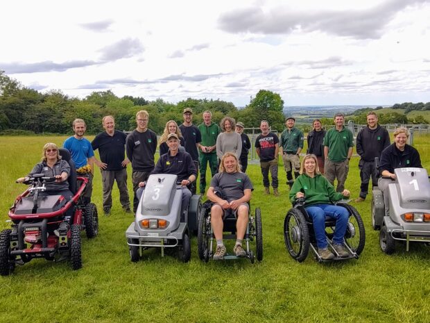 A group photo in a field. Some people are standing, others are seated in a variety of mobility aids and all terrain vehicles. All are smiling.