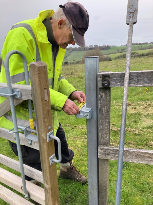 Man wearing a baseball cap and a hi-his jacket closely inspecting the latch on a gate in a field.