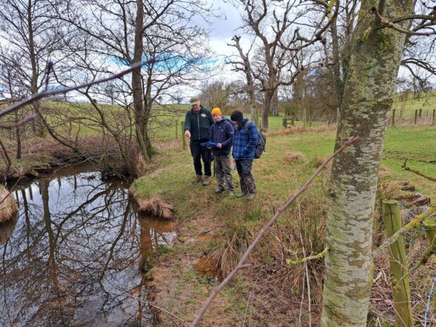 Three people in hiking gear stood on the bank of a river, looking at an iPad. One man is pointing at the river.
