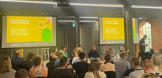 Kate Whitehead, Natural England Senior Officer for Children and Nature taking part in a roundtable discussion around Nature Connecting Schools. A panel of people sit facing an audience, in discussion, in front of two screens showing a title slide for the presentation.