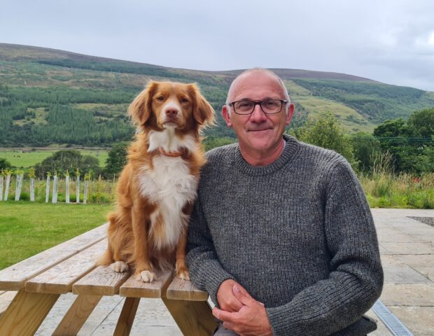 A man sits on an outdoor picnic table seat, facing the camera and smiling. His dog sits on the table top, next to him, also looking at the camera. In the background there are green hills, and cloudy sky.