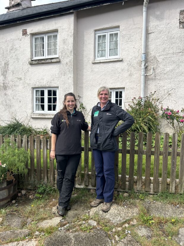 Two smiling women in hiking gear standing next to each other against a garden fence with a white building in the background.