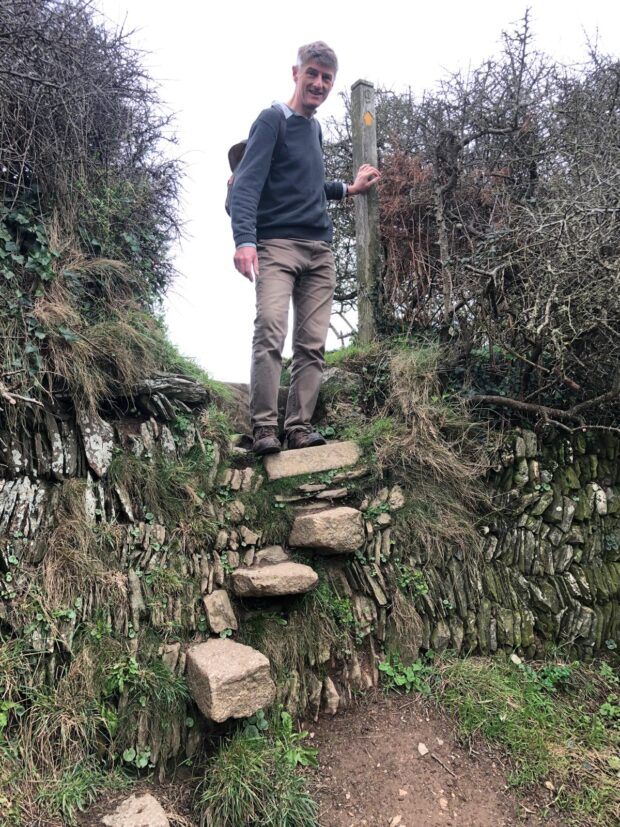 A man standing on a rocky stile next to a signpost with the national trails logo on it.