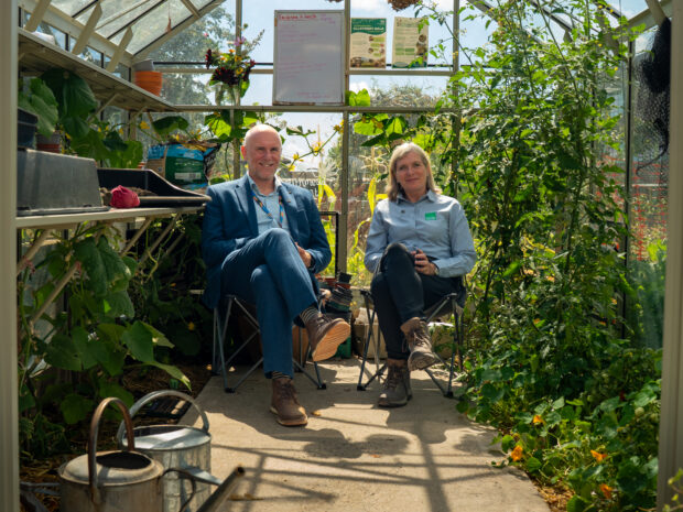 A man and a woman in formal work clothes with hiking boots sit in deck chairs in a greenhouse filled with plants, smiling and facing the camera.
