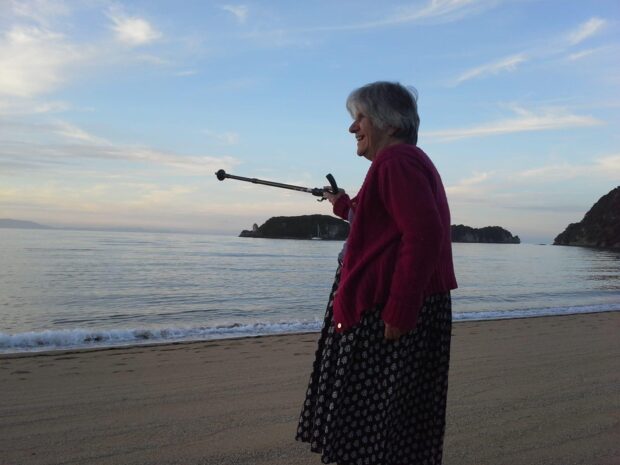 An older woman with a walking stick stands on the beach, smiling and pointing to the sea with her mobility aid.